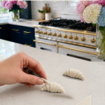Hand using a black-handled clay detailing tool to score horizontal layer lines into a raw white air dry clay croissant on a white quartz countertop, second refined clay croissant and an unrolled flat clay triangle visible nearby, pink peonies and white and blue hydrangeas in glass vases, navy blue kitchen cabinetry with brushed gold hardware and professional gold-knob range with white subway tile backsplash in background, step six of air dry clay croissant magnets tutorial refining the shape and defining pastry layers on jenniferdawn.ca
