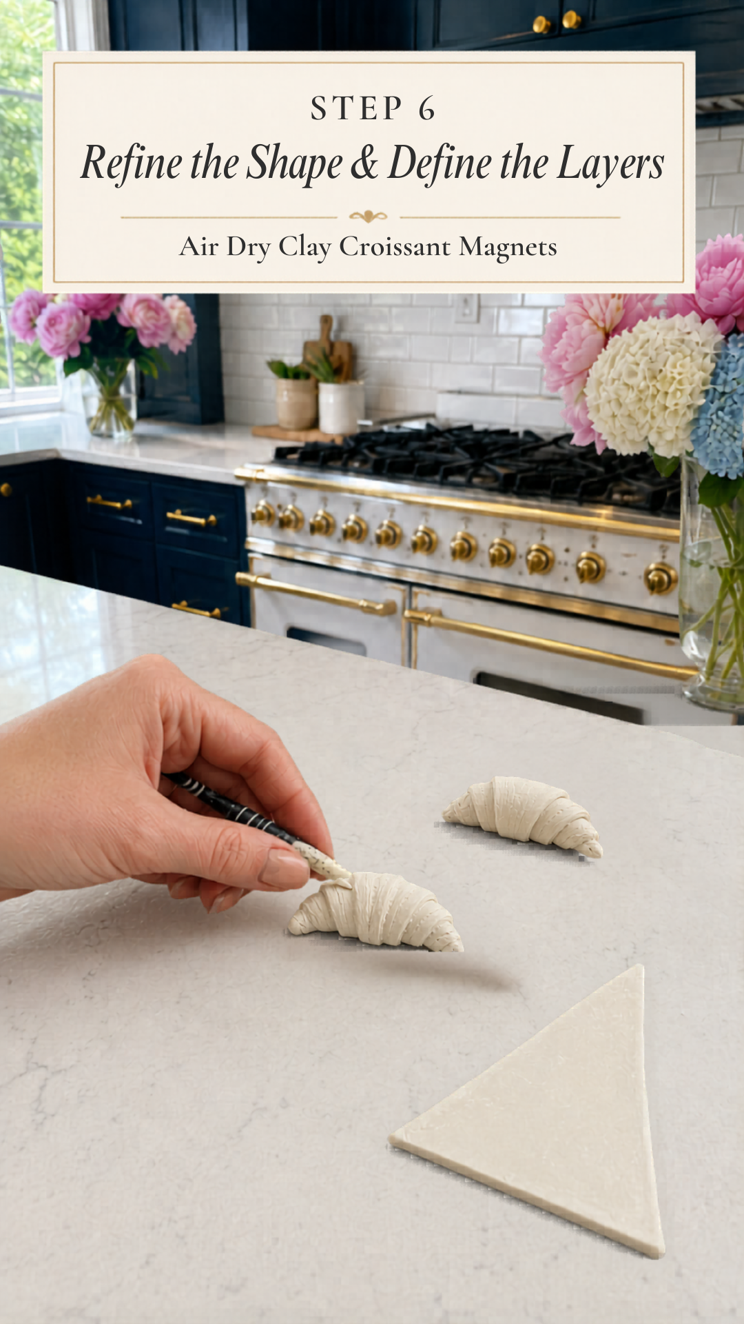 Hand using a black-handled clay detailing tool to score horizontal layer lines into a raw white air dry clay croissant on a white quartz countertop, second refined clay croissant and an unrolled flat clay triangle visible nearby, pink peonies and white and blue hydrangeas in glass vases, navy blue kitchen cabinetry with brushed gold hardware and professional gold-knob range with white subway tile backsplash in background, step six of air dry clay croissant magnets tutorial refining the shape and defining pastry layers on jenniferdawn.ca