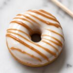 Overhead close-up of a single finished polymer clay donut magnet with a classic ivory glaze base and a thin irregular caramel-toned drizzle line across the top on a white marble surface with a toothpick resting beside it