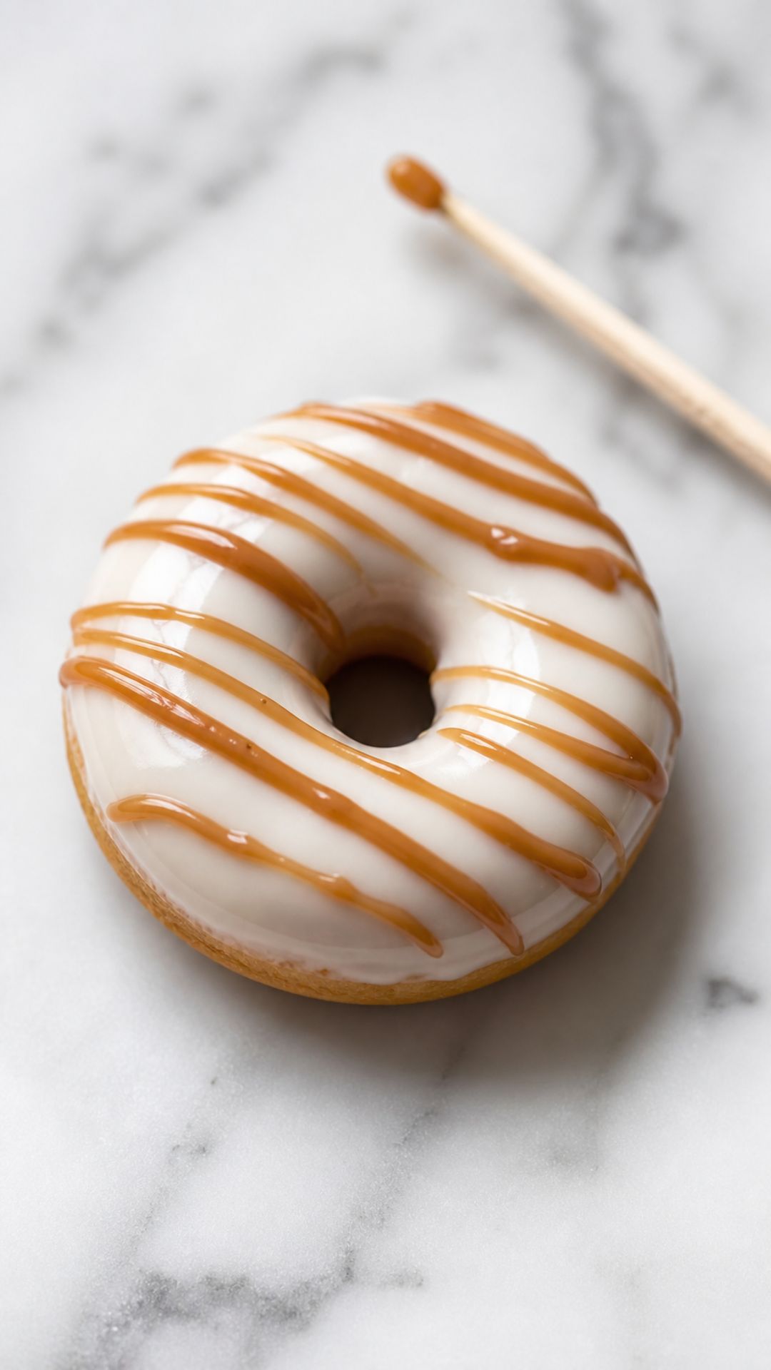 Overhead close-up of a single finished polymer clay donut magnet with a classic ivory glaze base and a thin irregular caramel-toned drizzle line across the top on a white marble surface with a toothpick resting beside it