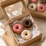 Handmade polymer clay donut magnets in pink, chocolate, and ivory glaze nestled in a kraft gift box with a clear window lid, tissue paper, and natural twine tag on a warm white surface