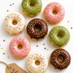 Overhead product display of twelve polymer clay donut fridge magnets in assorted glaze colors arranged on a white surface with a handwritten price tag reading 3 for dollar 15