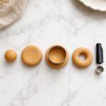 Overhead flat lay showing four stages of shaping a polymer clay donut base on white marble — clay ball, pressed disc, outer edge cut, and finished donut ring with a pen cap and circle cutter beside them