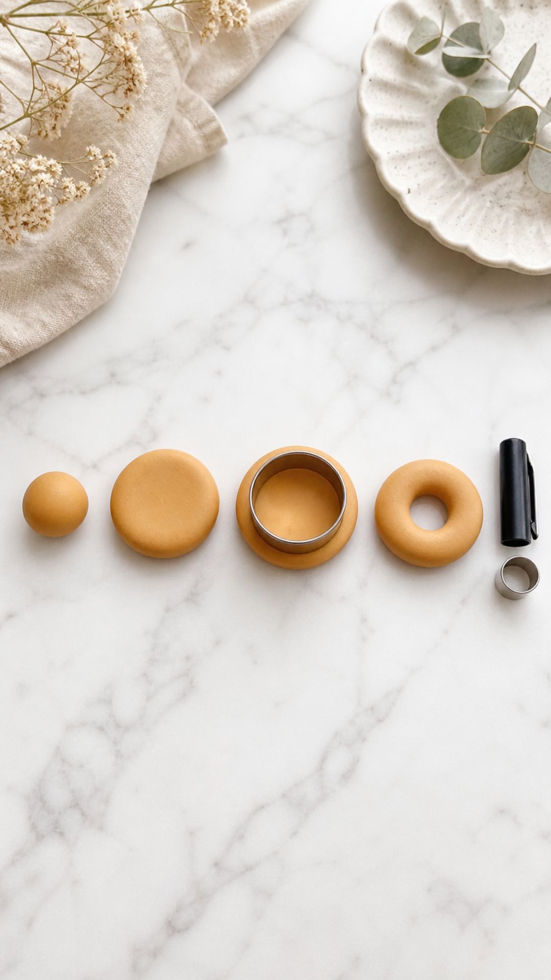 Overhead flat lay showing four stages of shaping a polymer clay donut base on white marble — clay ball, pressed disc, outer edge cut, and finished donut ring with a pen cap and circle cutter beside them