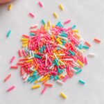 Close-up of tiny colorful handmade polymer clay sprinkles in pink, yellow, turquoise, white and coral scattered on a white marble surface with a fingertip visible for scale