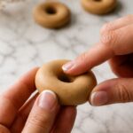 Close-up of a fingertip gently pressing down on the center top of a raw golden tan polymer clay donut ring to create a subtle dome shape on a white marble surface