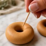 Close-up of a wooden toothpick being dragged very lightly across the domed surface of a raw golden tan polymer clay donut ring creating barely-visible texture marks on white marble