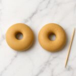 Overhead view of two raw golden tan polymer clay donut rings side by side on white marble — left donut smooth and untextured, right donut with subtle toothpick surface texture, wooden toothpick resting beside them