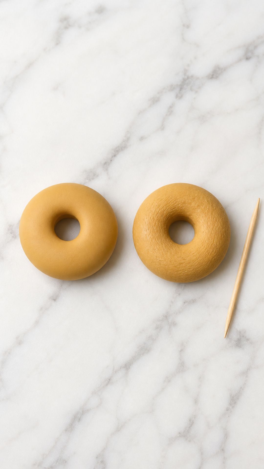 Overhead view of two raw golden tan polymer clay donut rings side by side on white marble — left donut smooth and untextured, right donut with subtle toothpick surface texture, wooden toothpick resting beside them