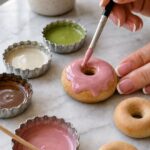 Fine detail brush applying pink liquid polymer clay glaze to a raw donut magnet on white marble with small mixing containers holding chocolate, ivory, and matcha tinted glazes nearby and a small succulent plant in the background