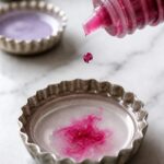 Extreme close-up of a small glass dropper bottle releasing a single drop of bright pink alcohol ink into a metal bottle cap container of translucent liquid polymer clay on a white marble surface with three other glaze mixing containers slightly out of focus in the background