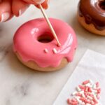 Close-up of a wooden toothpick carefully placing a single tiny coral polymer clay sprinkle onto the wet pink glazed surface of a polymer clay donut magnet on white marble with small piles of white and coral sprinkles on parchment nearby