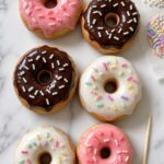 Overhead flat lay of six finished baked polymer clay donut fridge magnets in pink, chocolate, and ivory glaze with colorful sprinkles arranged in two rows on a white marble surface