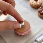 Close-up of a finger pressing a small silver neodymium disc magnet onto the flat bottom of a finished pink polymer clay donut magnet with a dot of E6000 adhesive visible on wax paper surface