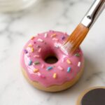 Fine soft brush applying a shiny gloss varnish coat over the pink glazed top of a finished baked polymer clay donut magnet on a white marble surface