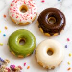 Overhead flat lay of four finished polymer clay donut fridge magnets in pink, chocolate, matcha green, and classic ivory glaze arranged on a white surface with dried flowers nearby