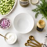 Overhead flatlay of low carb dill pickle salad in a white bowl surrounded by small bowls of shredded chicken, crispy bacon, diced cheddar, sliced avocado, and chopped hard-boiled eggs on a white quartz countertop with dark navy cabinetry in the corner.