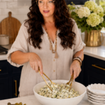 Woman with long dark hair tossing a large bowl of dill pickle salad with gold tongs in a luxury modern farmhouse kitchen with dark navy blue cabinetry, gold hardware, white subway tile, and white hydrangeas in a gold vase.