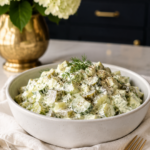 White speckled bowl of creamy low carb dill pickle salad garnished with fresh dill and cracked black pepper, placed on a white linen napkin with a gold fork, white hydrangeas in a gold vase and dark navy cabinetry in the background.