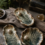 Three organic-shaped air dry clay incense holder dishes with detailed fern leaf impressions pressed into the clay, glazed in moody earthy tones of cream, forest green, and rust brown, each holding a small spherical incense stick holder, styled on a weathered driftwood plank with crystals, botanical books, and fresh fern fronds in a dark woodland flat lay.