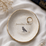 Overhead flat lay of a cream speckled air dry clay ring dish with irregular organic edges, gold rim detailing, a gold ring with crystal stone resting inside, stamped with the name Jennifer Dawn and a small raven illustration, surrounded by white linen fabric and dried baby's breath flowers.