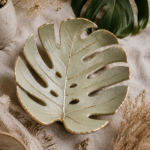 Overhead flat lay of a large air dry clay monstera leaf dish with detailed leaf vein impressions throughout, painted in sage green with brushed gold edges along the rim and veins, styled on a warm beige linen cloth with a real monstera leaf, dried pampas grass, baby's breath in a speckled ceramic vase, and a partial view of a jewelry dish in the lower left corner.