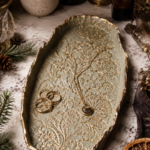 Overhead angled view of a large oval air dry clay jewelry tray painted sage green with intricate gold botanical branch and baby's breath impressions pressed across the entire surface and gold-leafed irregular edges, holding three gold stacking rings and a gold chain necklace with a labradorite pendant, styled on a white marble surface with fresh pine sprigs, pine cones, crystals, dried hydrangeas, baby's breath, amber apothecary bottles, and a lit candle in a moody witchy flat lay.