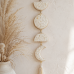 White air dry clay moon phase wall hanging with five moon shapes in varying phases — crescent, quarter, full, and waning — embossed with gold constellation patterns and connected by small gold chain links, ending in a long cream and gold macrame tassel, hung on a warm plaster wall beside a vase of dried pampas grass.