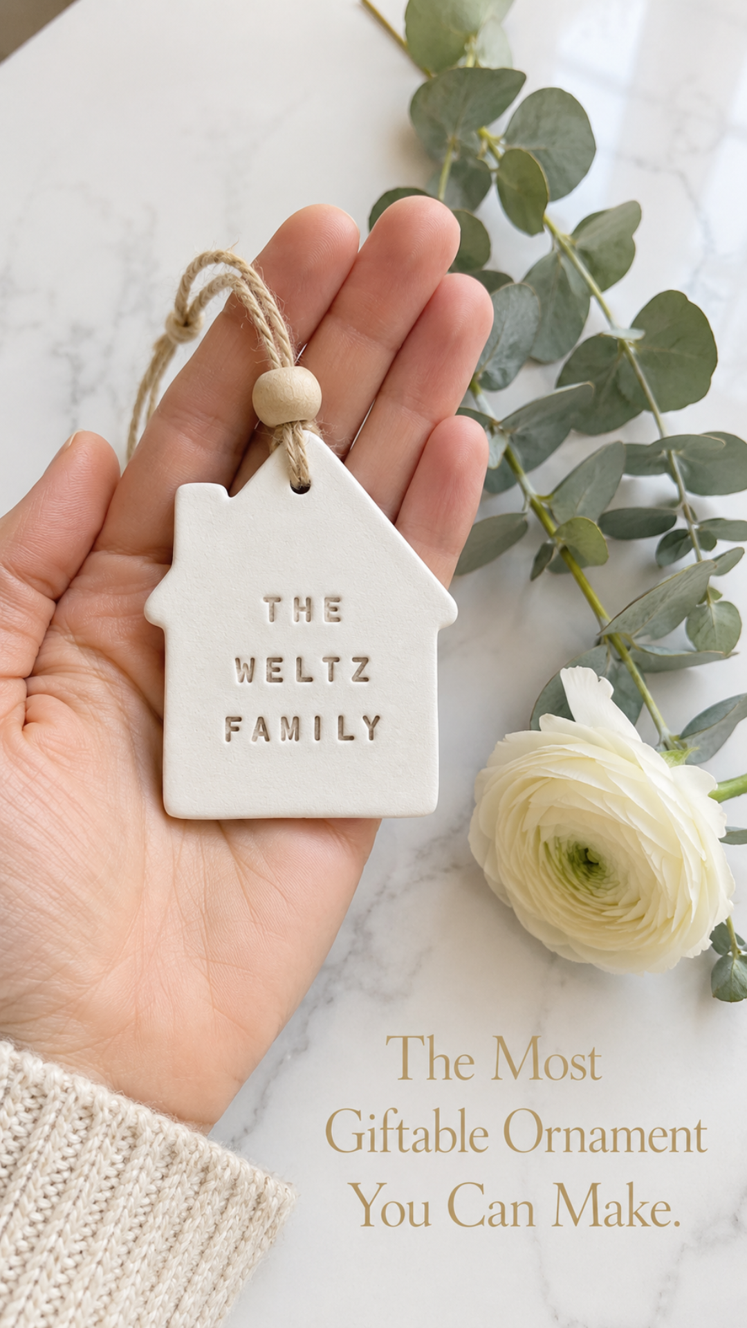 Close-up of an open palm holding a smooth white air dry clay house-shaped Christmas ornament stamped with the words THE WELTZ FAMILY in clean block letters. Natural jute twine loop with a small round wooden bead at the top of the ornament. A fresh eucalyptus stem and a single cream ranunculus bloom rest beside the hand on a white marble surface. Text overlay in warm gold serif reads The Most Giftable Ornament You Can Make.