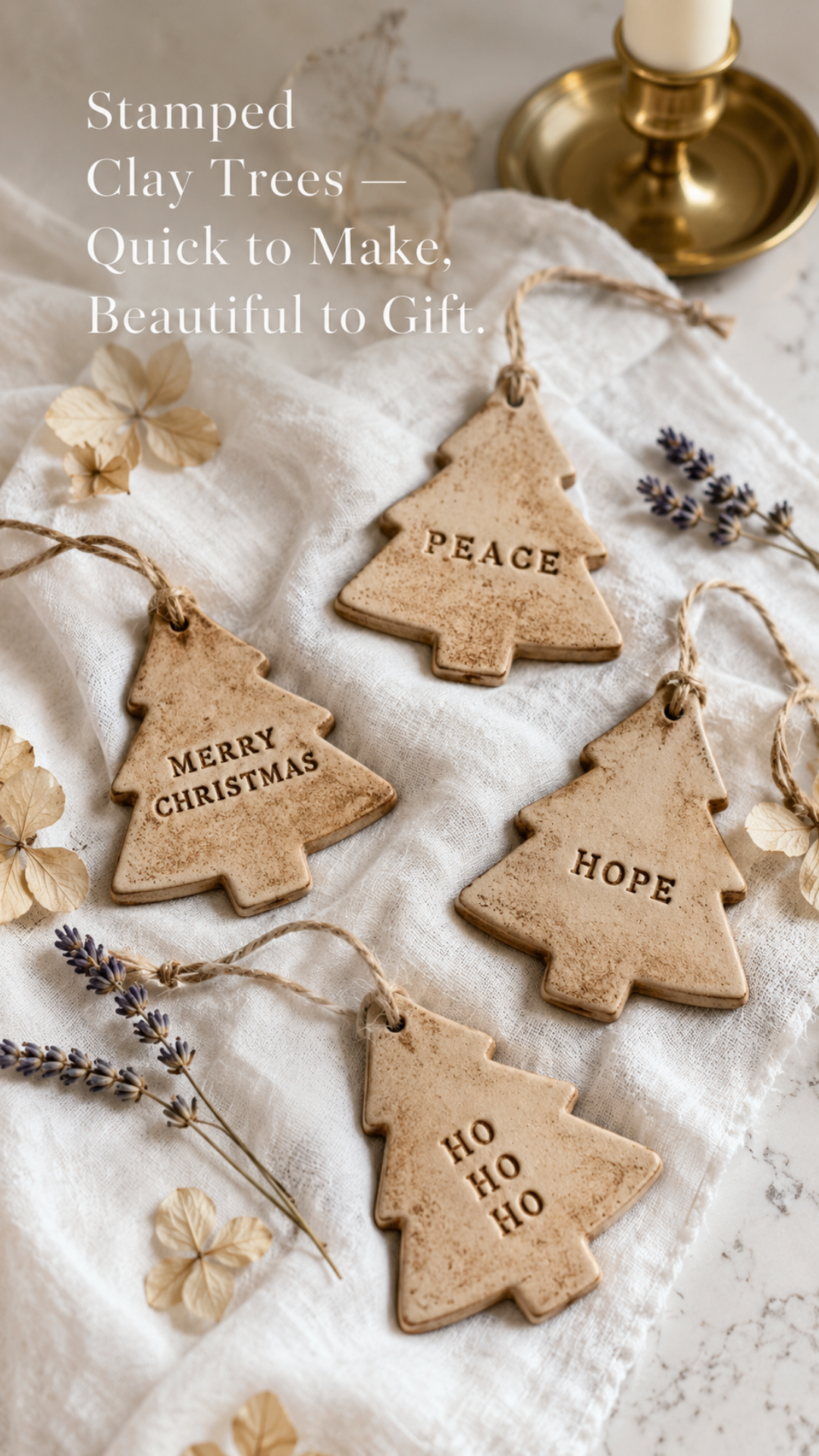 Styled flat lay of four handmade air dry clay Christmas tree ornaments on a white linen cloth. Each tree shape is stamped with a different word — PEACE, MERRY CHRISTMAS, HOPE, and HO HO HO — in clean block letter stamps with a warm antiqued brown wash finish. Natural jute twine loops at the top of each ornament. Dried hydrangea petals and sprigs of lavender are scattered around the ornaments with a brushed gold taper candleholder visible in the upper right corner. Text overlay reads Stamped Clay Trees Quick to Make Beautiful to Gift.