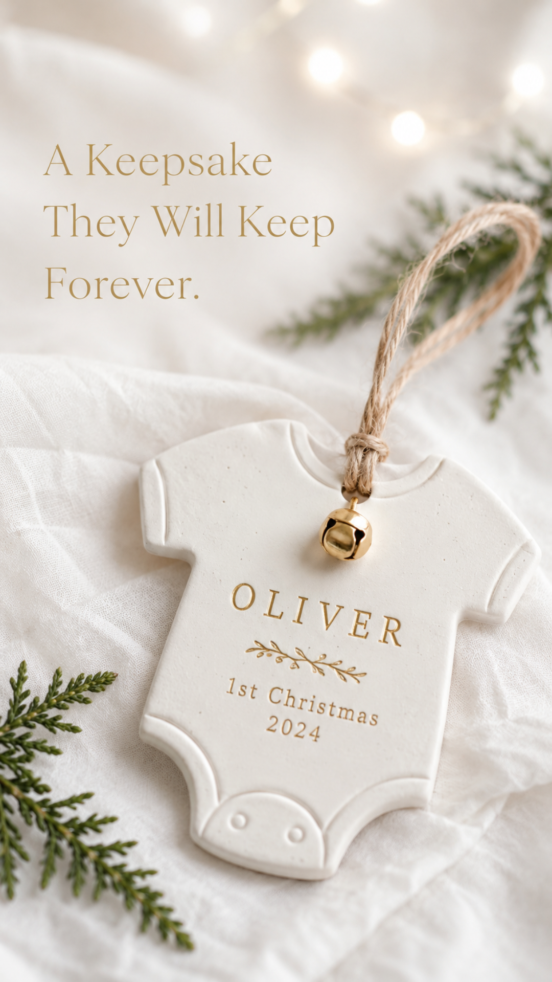 Close-up macro shot of a smooth white air dry clay onesie-shaped Christmas ornament on white crinkled linen. The ornament is stamped with the name OLIVER and the words 1st Christmas 2024 in serif letters with gold paint worked into the stamped impressions. A small gold jingle bell hangs at the collar with a natural jute twine loop above. Small sprigs of fresh cedar rest beside the ornament with warm fairy light bokeh glowing softly in the background. Text overlay in warm gold serif reads A Keepsake They Will Keep Forever.