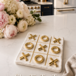 White clay tic tac toe board with brushed gold X and O pieces on a white quartz countertop, styled with pink peonies, hydrangeas, and ranunculus, dark navy cabinetry and gold appliances visible in the background.