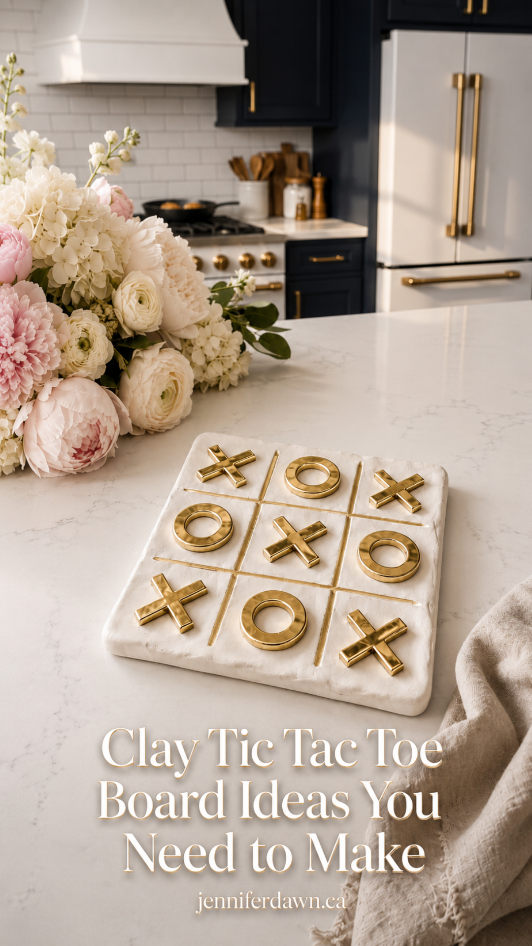 White clay tic tac toe board with brushed gold X and O pieces on a white quartz countertop, styled with pink peonies, hydrangeas, and ranunculus, dark navy cabinetry and gold appliances visible in the background.