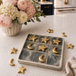 Dark marbled clay tic tac toe board with gold star and crescent moon pieces scattered around it, styled on a white quartz countertop with a pink and cream floral arrangement, navy cabinetry and gold refrigerator in the background.