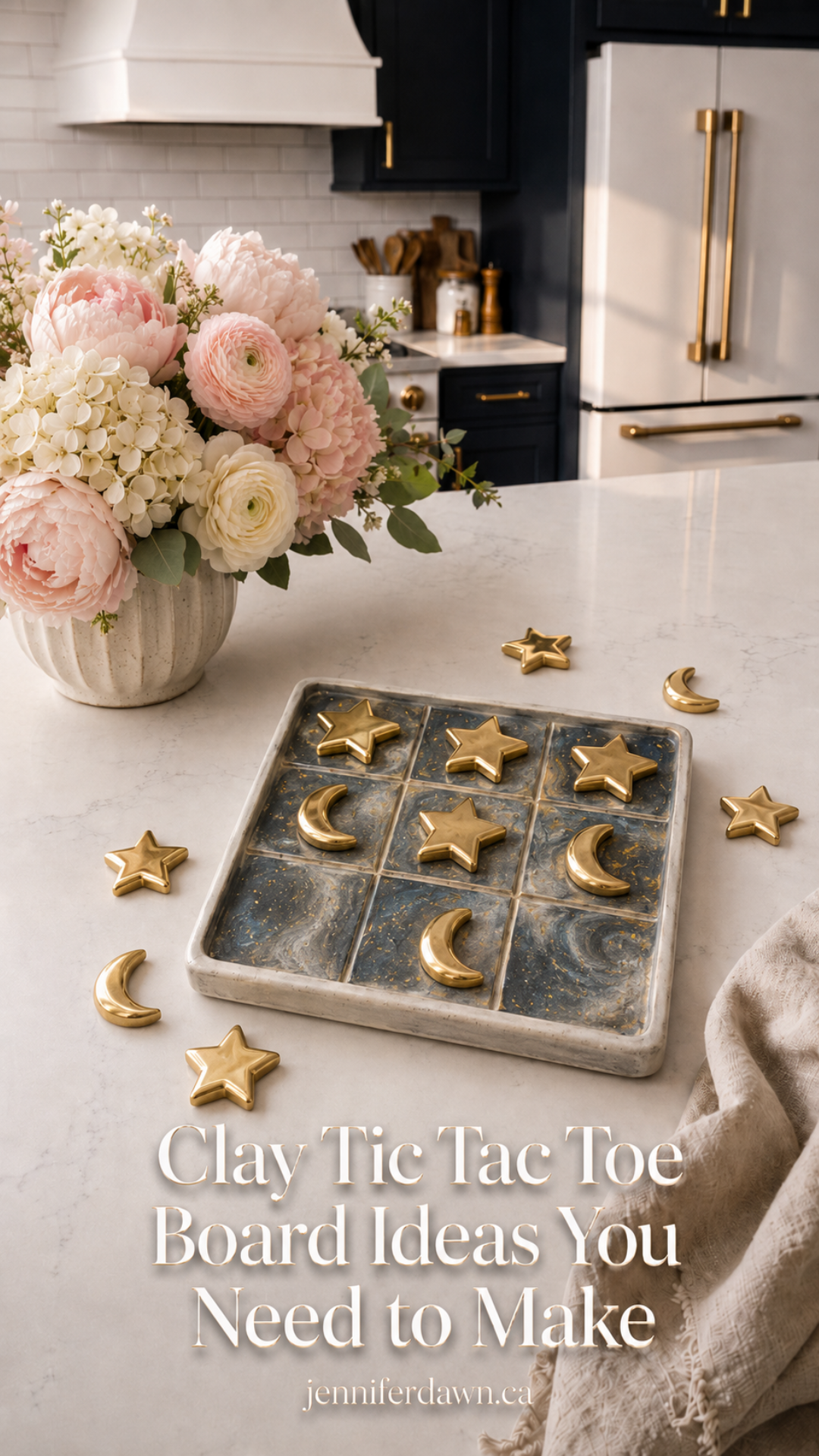 Dark marbled clay tic tac toe board with gold star and crescent moon pieces scattered around it, styled on a white quartz countertop with a pink and cream floral arrangement, navy cabinetry and gold refrigerator in the background.