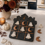 Black clay haunted house-shaped tic tac toe board with white ghost pieces and copper crescent moon pieces on a white quartz countertop, styled with a deep autumn floral arrangement, white pumpkin, and lit amber candle, navy and gold kitchen in background.