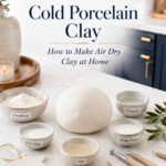 Cold porcelain clay ingredients labeled cornflour, oil, vinegar, and white paint arranged on a white marble countertop with a smooth ball of homemade air dry clay and finished decorative clay pieces including a clay cottage, sculpted rose, and embossed ornament in a navy and gold kitchen.