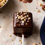 Hand sprinkling chopped hazelnuts onto a low carb chocolate covered marshmallow pop, keto candy recipe