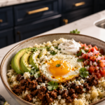 Speckled ceramic bowl filled with seasoned cauliflower rice, crumbled Mexican chorizo, a fried egg with a runny yolk, sliced avocado, fresh pico de gallo, sour cream, and crumbled cotija cheese on a white marble countertop with dark navy cabinetry and brushed gold hardware softly blurred in the background.