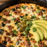 Cast iron skillet filled with a golden baked keto chorizo and poblano frittata topped with salsa verde, fanned avocado slices, and fresh cilantro, styled on a linen napkin with a bowl of salsa verde and stacked white plates softly blurred in the background.