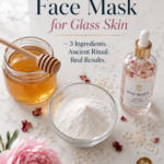 Overhead flat lay of rice flour face mask ingredients on white marble countertop with dark navy cabinetry — glass bowl of rice flour, honey jar with wooden dipper, rose water bottle, pink peonies, scattered rose petals, and gold measuring spoon.