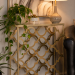 Brass quatrefoil patterned console table with a glass top in a warm entryway, styled with a trailing pothos plant, a small succulent, and a ribbed ceramic table lamp against a marbled mirror.