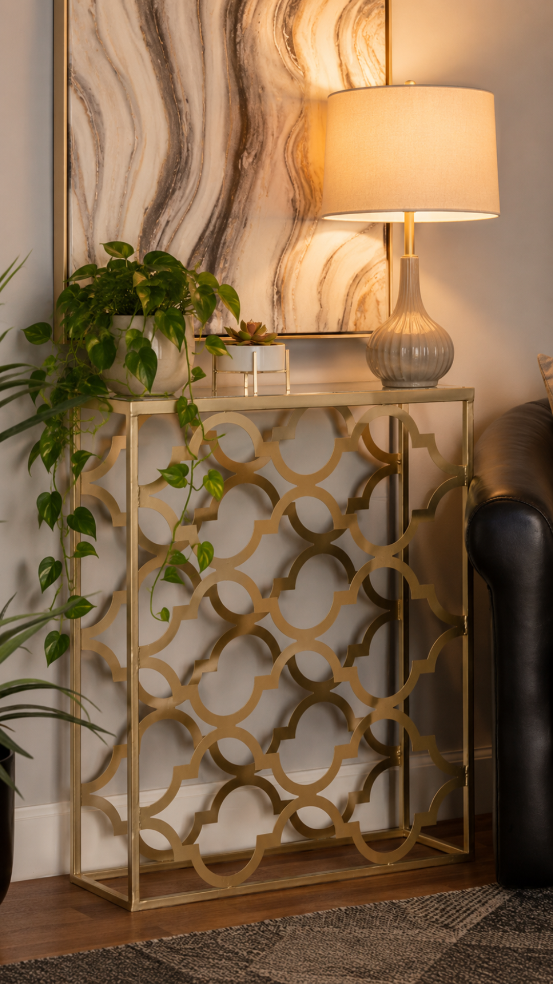 Brass quatrefoil patterned console table with a glass top in a warm entryway, styled with a trailing pothos plant, a small succulent, and a ribbed ceramic table lamp against a marbled mirror.