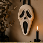 A cream air-dry clay ghost face wall decor piece with elongated oval shape, wide dark eyes, and open mouth, hanging from a black satin ribbon with dried eucalyptus and a lit black taper candle nearby