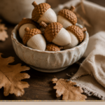 A small ceramic bowl filled with textured air-dry clay acorns with warm brown caps and cream nut bodies, styled with dried oak leaves and a linen napkin on a dark wood surface