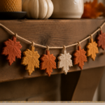 An air-dry clay maple leaf garland in warm red, burnt orange, mustard, and cream strung on jute twine, draped across a wooden mantel with a beeswax candle, small white pumpkin, and vase of dried autumn stems