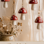 Five red-and-white spotted air-dry clay mushroom ornaments hanging on jute twine in front of a cream linen curtain with a vase of dried wildflowers below