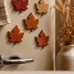 Five air-dry clay maple leaf magnets in warm red, burnt orange, and gold displayed on a cream vintage-style refrigerator with a striped linen tea towel and a vase of dried grasses