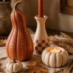 Coordinated air-dry clay fall decor trio featuring a tall terracotta sculpted gourd with hand-painted cream and gold dot detail, a cream candlestick with terracotta checkered base holding a burnt-orange taper candle, and a small pumpkin-shaped tea light holder with a glowing tea light, styled on a cream linen runner with dried leaves and wheat