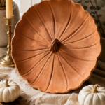 Large air-dry clay decorative plate in warm terracotta with curved ridge lines radiating out from a twisted knotted center stem, edges lifting gently upward, displayed leaning against dried pampas grass and eucalyptus with a brass candlestick, mini white pumpkins, and a cream fringed linen runner